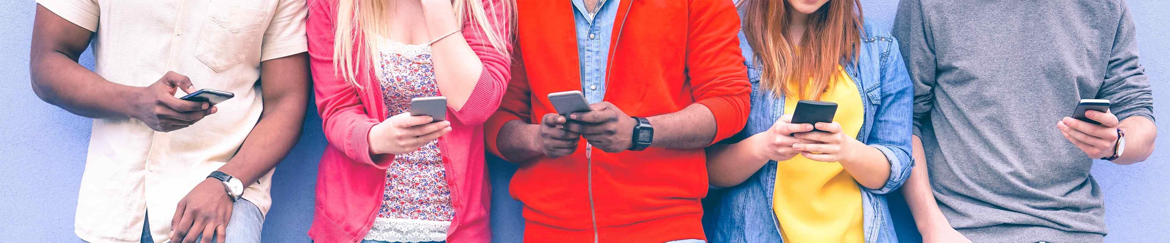 Five teens in brightly colored shirts standing in a row against a blue wall. All are holding their mobile phones and using social media.